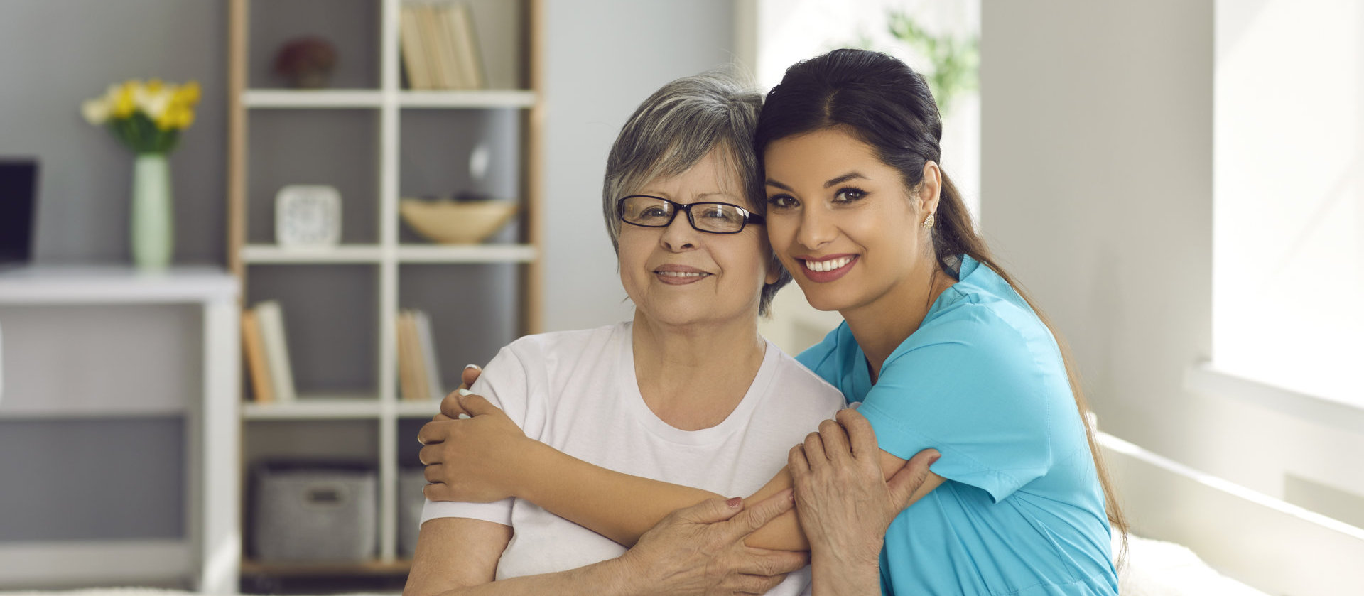 Supportive carer with her elderly patient smiling and looking at camera sitting on bed in retirement home